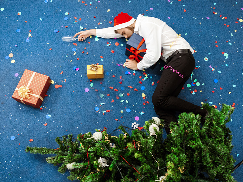 Person mit Weihnachtsmütze liegt zwischen Geschenken, Konfetti und einem umgestürzten Weihnachtsbaum.